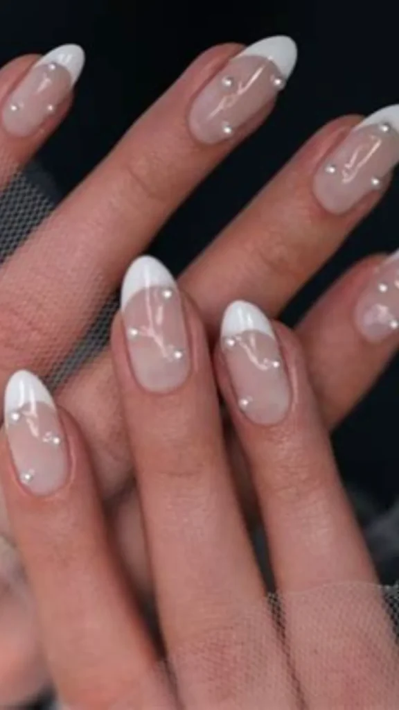 Close-up of a bridal manicure showing almond-shaped nails with classic white French tips on a nude pink base. Each nail is intricately embellished with scattered tiny white pearls against a dark background with sheer white bridal tulle fabric.
