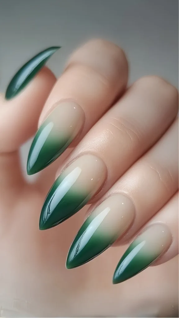 Close-up of a hand showcasing a seamless green ombré manicure on long, sharp stiletto-shaped nails in the file 1_4.webp. The design features a smooth gradient fade, transitioning beautifully from a glossy neutral nude shade at the cuticle base into a rich, deep emerald green toward the sharp tips.