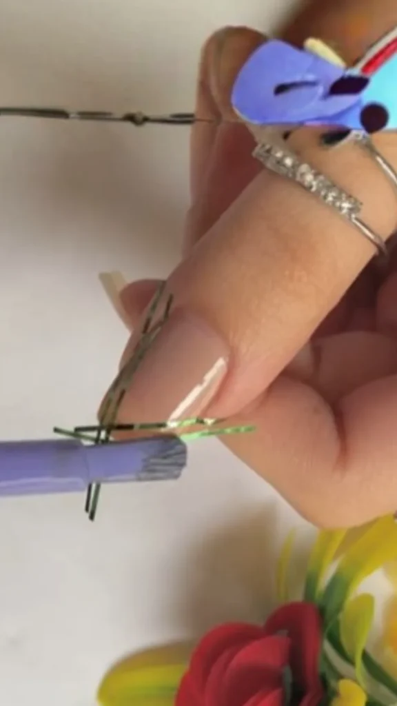 Close-up of a person's manicured fingernail being painted with light purple nail polish. A thin green hair elastic is wrapped vertically around the nail to act as a DIY stencil for creating clean, parallel striping lines.