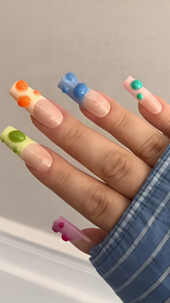 Close-up of a hand featuring a playful, multi-colored 3D French manicure on long, square-shaped nails. Each nail pairs a neutral nude base with a pastel-colored tip—including lime green, orange, blue, pink, and purple. The tips are uniquely decorated with raised, glossy 3D droplets in coordinating darker shades. The hand is pulled slightly into a blue striped button-down shirt sleeve.