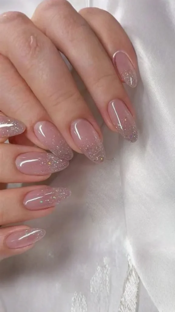 Close-up of a bridal manicure featuring long, glossy, almond-shaped nails. The design has a sheer nude-pink base that fades into fine silver and iridescent glitter concentrated at the tips, creating a subtle ombré sparkle effect against a soft white satin fabric background.