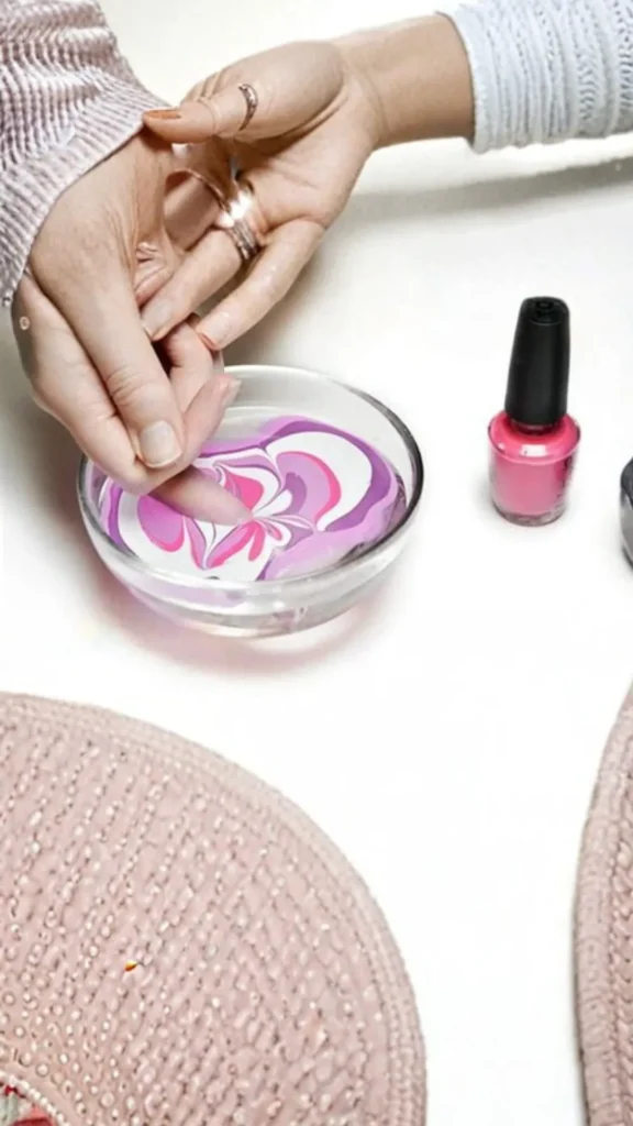 High-angle view of a person dipping their fingernail into a small glass bowl filled with water for water marble nail art. Swirls of pink, purple, and white nail polish float on the surface of the water, creating a floral pattern. A bottle of pink nail polish sits on the white tabletop next to the bowl.