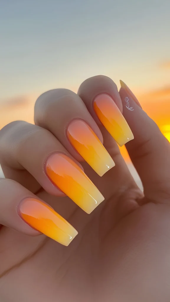 Close-up of a hand featuring a vibrant summer ombré manicure on long, square-shaped nails. The design showcases a bright gradient fade shifting seamlessly from a soft peach at the base into a rich orange and ending in a vivid yellow at the tips, set against a warm sunset sky background.