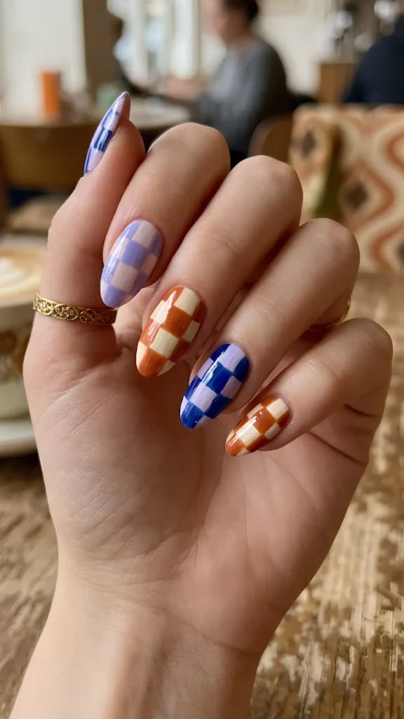 Close-up of a hand showcasing a playful, retro checkerboard manicure on long, oval almond-shaped nails. The design features a glossy, high-shine finish with alternating color schemes on each nail: the index and ring fingers feature a purple and cobalt blue grid, while the middle and pinky fingers display a burnt orange and cream grid. The hand wears a textured gold band ring and rests over a rustic wooden table with a soft-focus cafe background.
