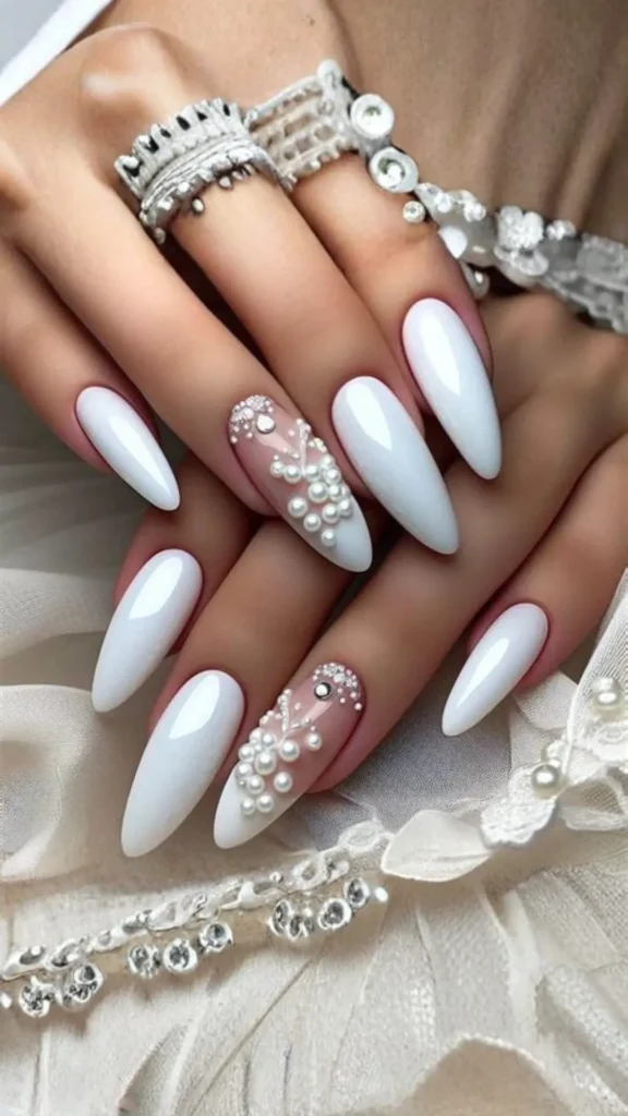 Close-up of two hands showing a glamorous bridal manicure with long, glossy, almond-shaped white nails. The ring fingers feature a sheer nude accent nail intricately embellished with clusters of various-sized white pearls and tiny clear rhinestones resembling delicate vines. The hands rest on elegant white lace and textured fabrics.