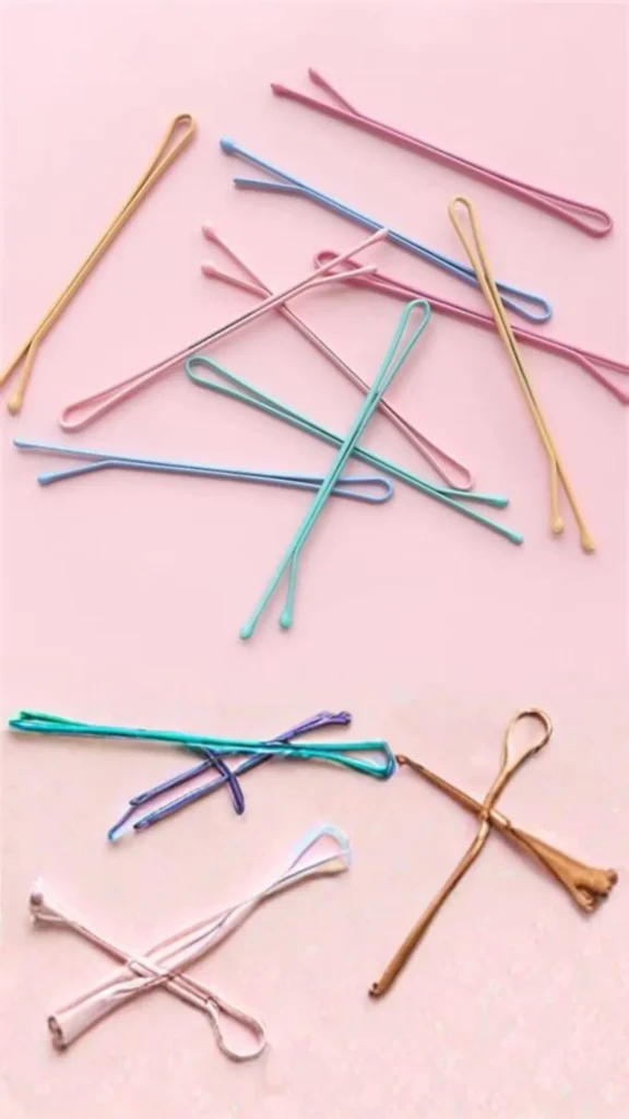 Top-down view of several bobby pins in various pastel shades like pink, blue, yellow, and green scattered across a solid pink background. The image highlights the rounded, bulbous tips and smooth curved loops of the pins, showcasing them as everyday items that can double as DIY tools.