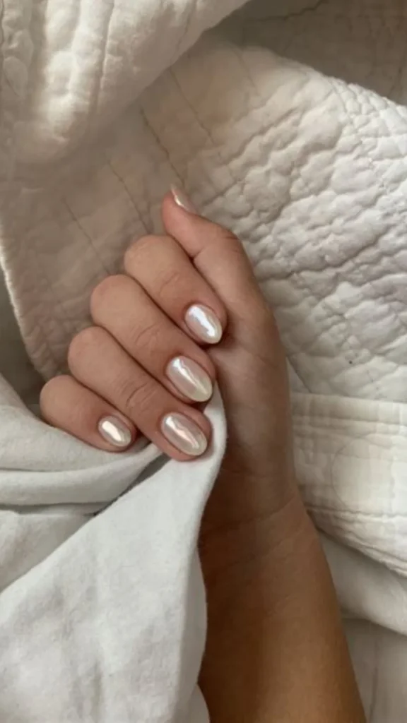 Close-up of a hand resting against a soft, textured white quilted blanket, showcasing an elegant "glazed donut" style manicure. The short, oval-shaped nails feature a pearlescent white chrome finish that gives off a luminous, iridescent glow.