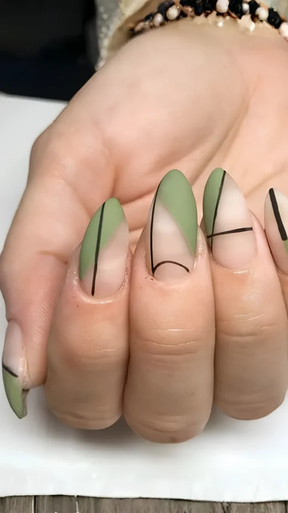 Close-up of a hand displaying a minimalist, geometric manicure on long, almond-shaped nails in the file 6_3.webp. The design features a smooth matte finish over a sheer nude base, creatively styled with sharp blocks of sage green polish in abstract triangular sections. The negative space and color blocks are defined by ultra-fine, crisp black lines forming intersecting stripes and a delicate curve near the cuticle.