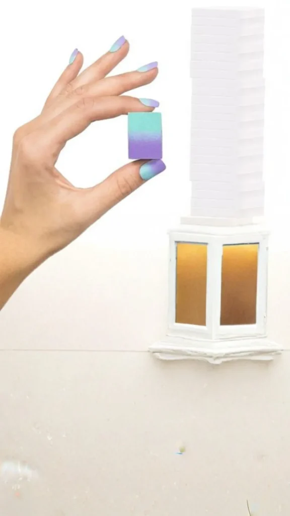 A person's hand with a matching teal and purple gradient manicure holds up a small rectangular makeup sponge painted with the same teal and purple ombré effect. In the background, a tall stack of clean, white makeup sponges sits next to a small white decorative structure.