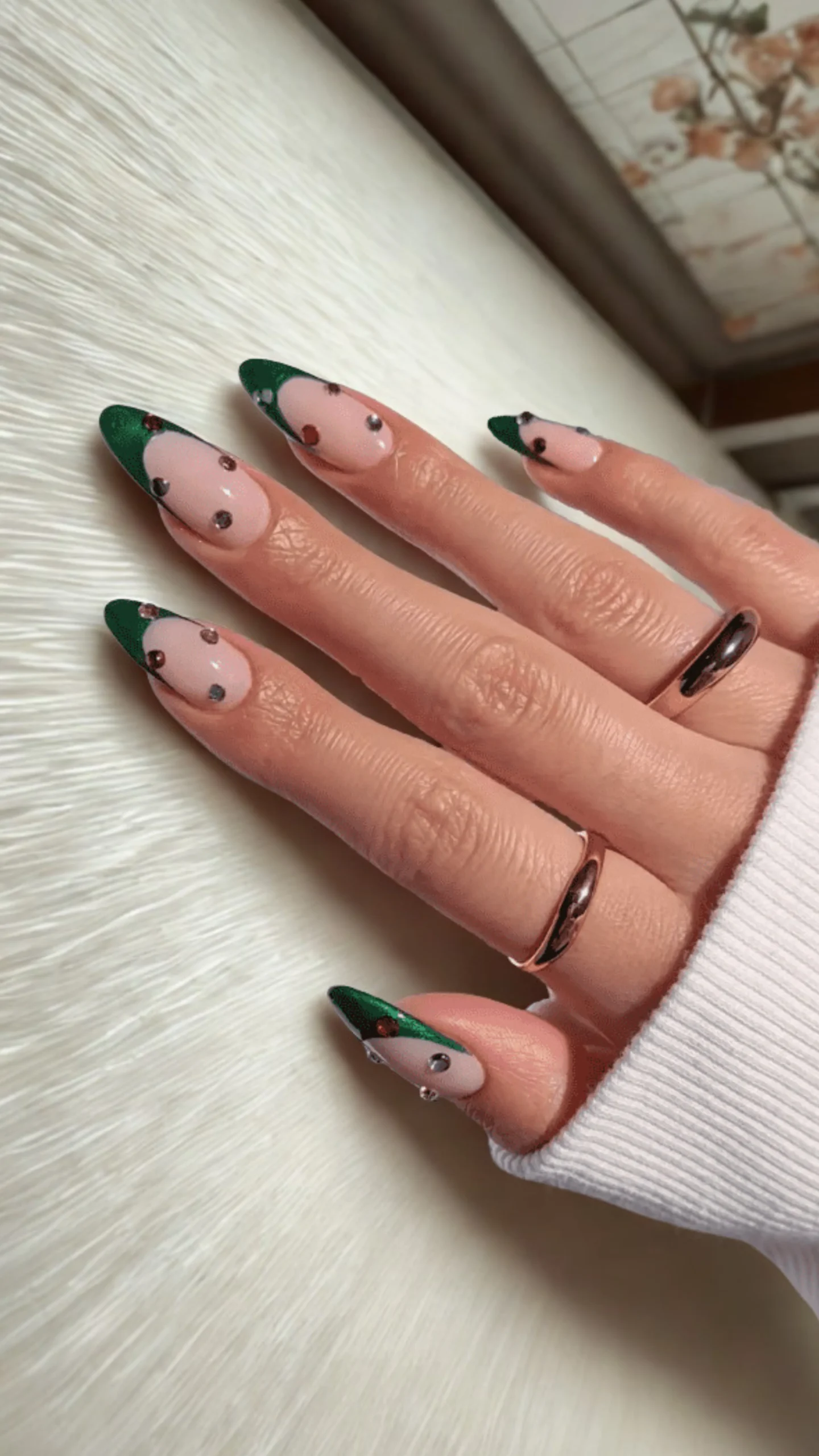 Close-up of a hand with long, almond-shaped nails displaying an elegant French manicure. The design features a smooth, nude-pink base paired with deep emerald green tips. The nails are further embellished with small, scattered rhinestones in clear, red, and dark tones. Two simple gold bands adorn the fingers against a white textured fabric background.