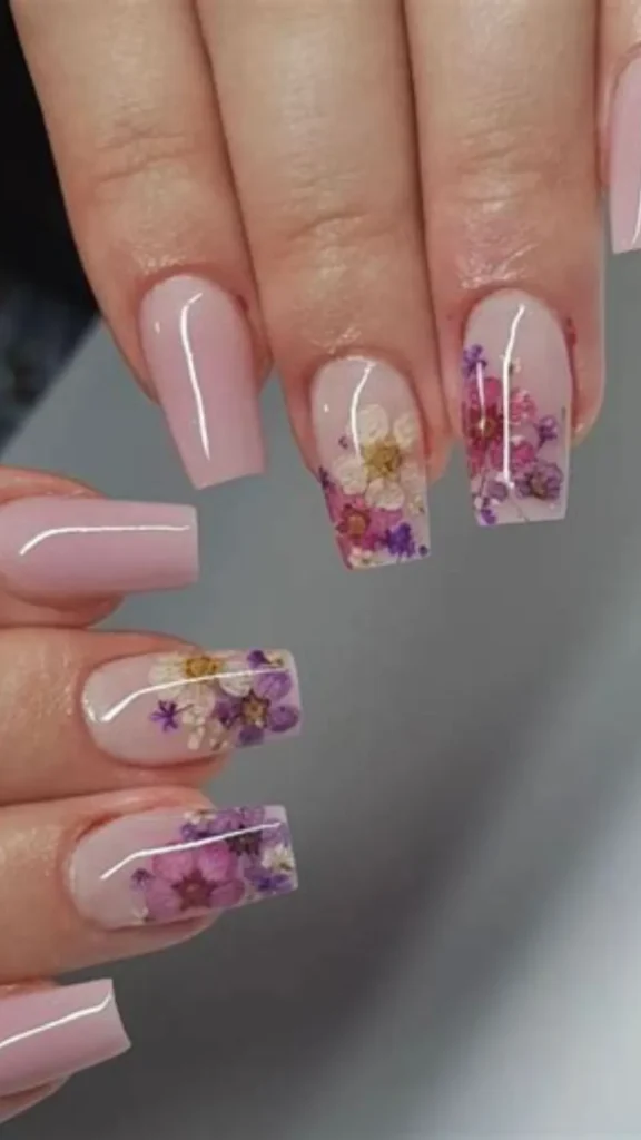 Close-up of hands showcasing an elegant bridal manicure with medium-length, glossy, square-shaped nails. Most of the nails feature a solid pastel pink coat, while select accent nails display an intricate design of colorful real dried flowers in shades of purple, pink, and white embedded under a clear, glossy top coat.