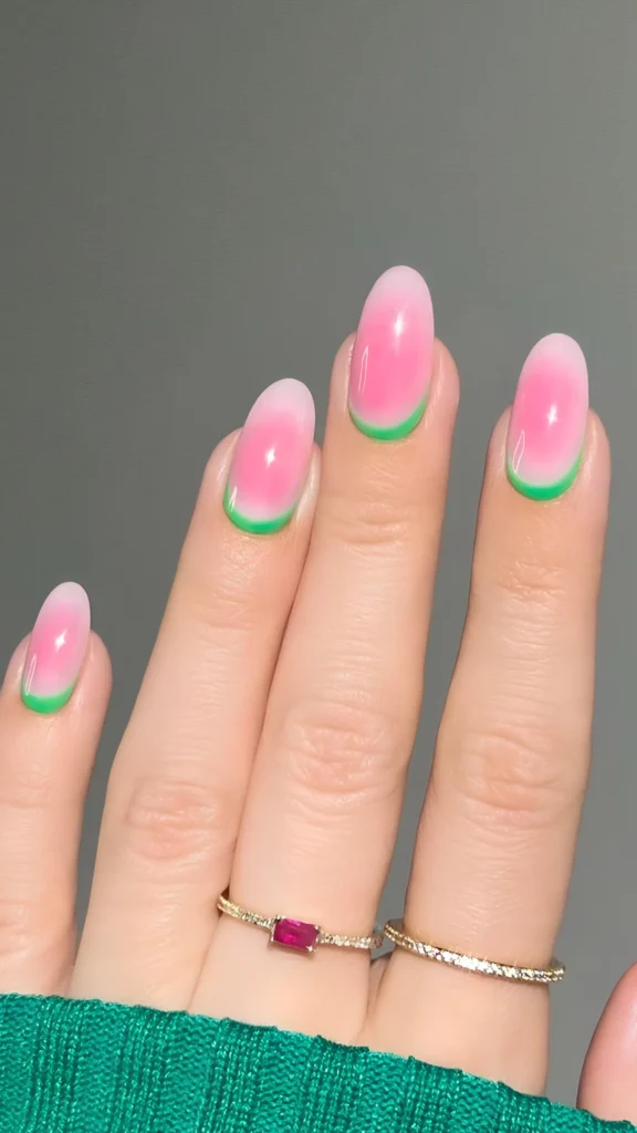 Close-up of a hand with medium-length, oval-shaped nails showcasing a creative reverse French manicure. The design features a vibrant pink aura or ombré base coat that fades beautifully into a milky-white edge, contrasted by a crisp, bright green crescent line tracing along the cuticle of each nail. The hand wears a ruby and a diamond ring, resting against a rich green knit fabric.