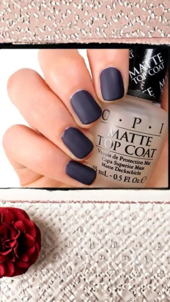 Close-up of a hand showcasing a smooth, matte dark navy blue manicure. The fingers are curled around a frosted glass bottle of OPI Matte Top Coat. A textured white background with a dark red flower is visible at the bottom.