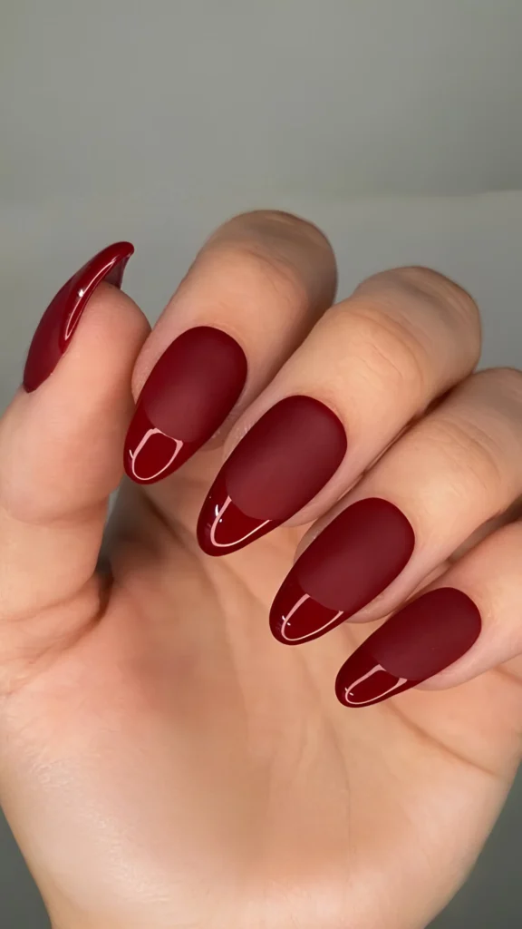 Close-up of a hand showcasing a dramatic French manicure on long, oval-shaped nails. The design features a deep burgundy red palette where the main body of each nail has a smooth, non-reflective matte finish, contrasted by ultra-glossy, shining burgundy red tips.