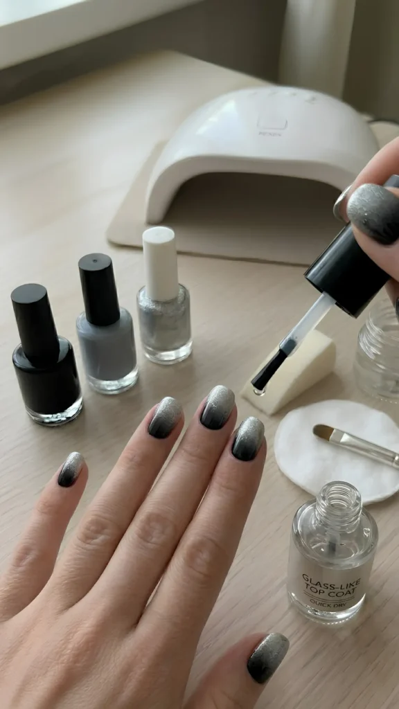 Close-up of a person's hand receiving a top coat over a black-to-silver ombré manicure on short, rounded nails over a light wooden table setup. The right hand holds a nail polish brush with a clear drop of top coat, hovering it over the index finger to seal the seamless gradient transition of black, gray, and sparkling silver glitter. In the foreground, an open bottle labeled "GLASS-LIKE TOP COAT QUICK DRY" sits open. In the background, bottles of black, gray, and silver glitter polish are lined up next to a white UV/LED nail lamp, a makeup sponge, and a clean-up brush resting on a cotton round.