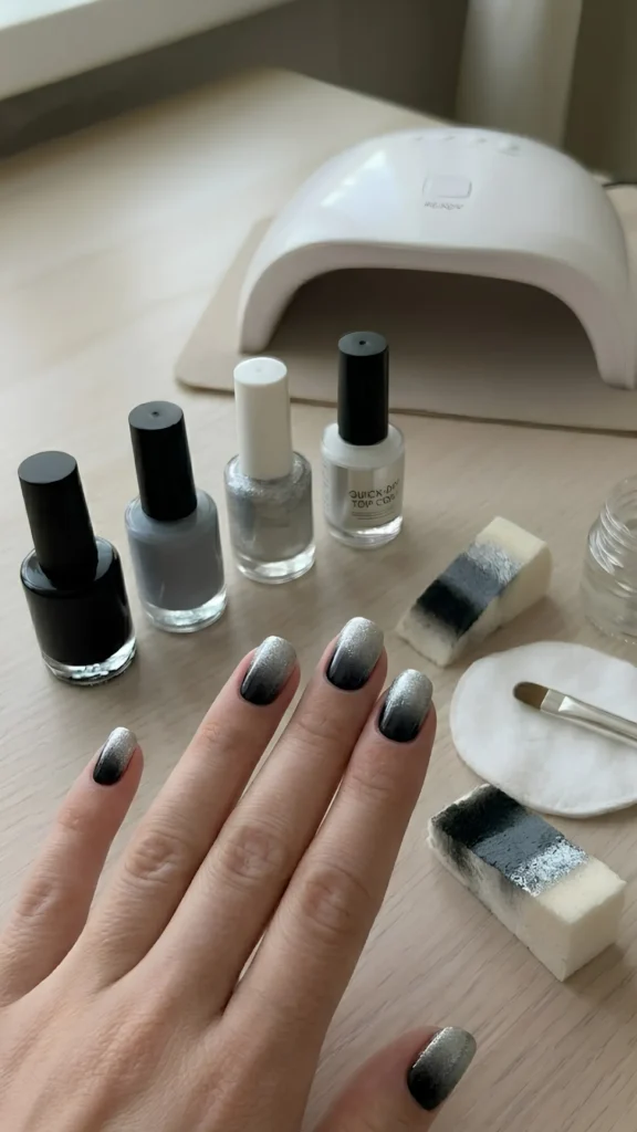Close-up of a person's hand showcasing an edgy and chic black-to-silver ombré manicure on short, rounded nails over a light wooden table setup. Each nail features a smooth, seamless gradient transition starting with a solid black base at the cuticle, blending into a medium gray in the middle, and ending with a sparkling metallic silver glitter at the tip. In the background, bottles of black, gray, and silver glitter polish are lined up alongside a quick-dry top coat, a white UV/LED nail lamp, a clean-up brush resting on a cotton round, and two rectangular makeup sponges stained with the black, gray, and silver ombré pattern.