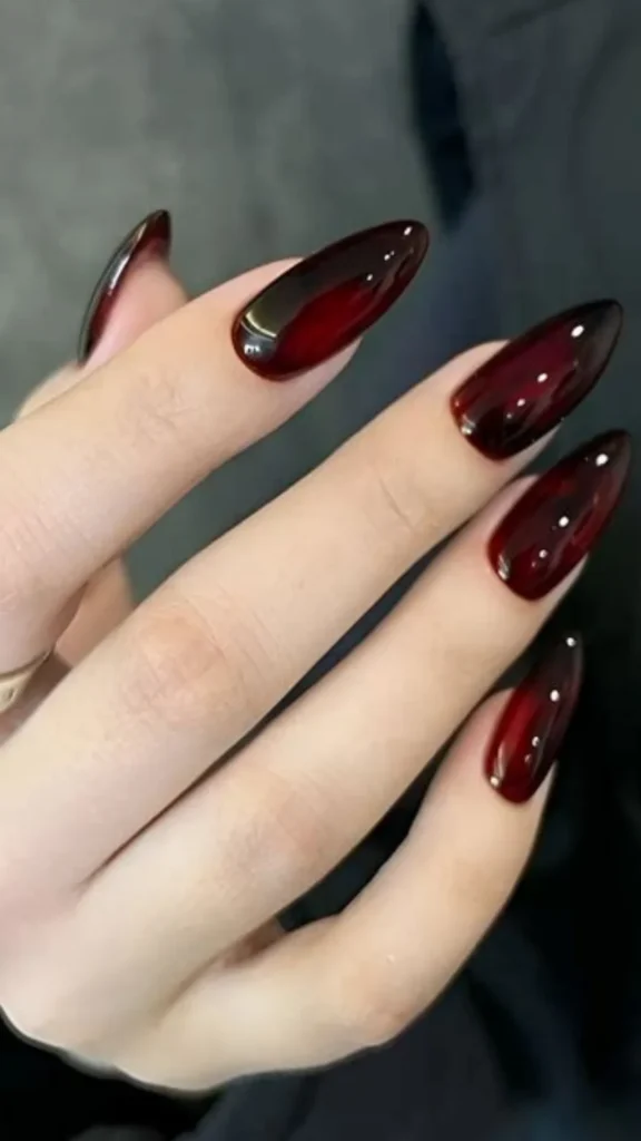 A close-up of a hand showcasing long, elegant almond-shaped nails. The nails feature a rich, high-gloss dark cherry red jelly polish with an ultra-shiny, translucent finish that creates a deep, multi-dimensional glass-like look.