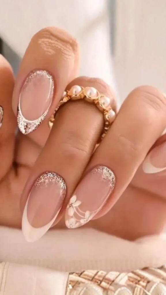 A close-up of a hand with elegant, almond-shaped nails showcasing a modern bridal French manicure. The nails feature clean white tips, a touch of fine champagne gold glitter at the cuticles, and a delicate hand-painted white floral accent on the ring finger.