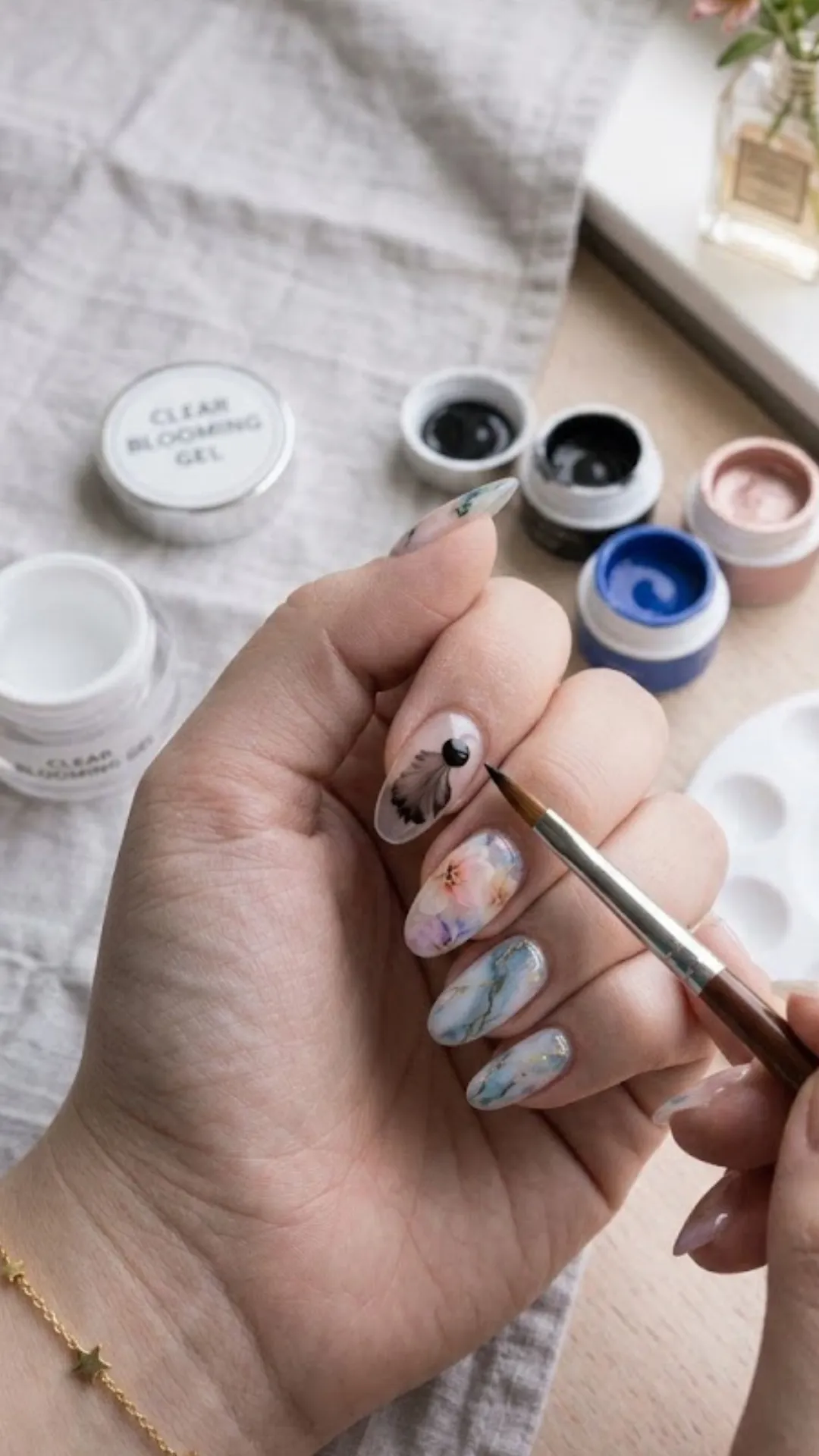 A close-up of a hand with a completed blooming gel manicure featuring delicate smoky watercolor textures and marble effects, while a nail tech uses a fine detail brush to refine a black watercolor design on the index finger.