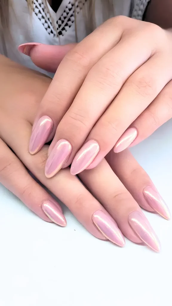 A close-up of hands featuring long, almond-shaped manicured nails painted in a soft blush pink shade with a highly reflective, iridescent chrome glaze overlay that catches the light.