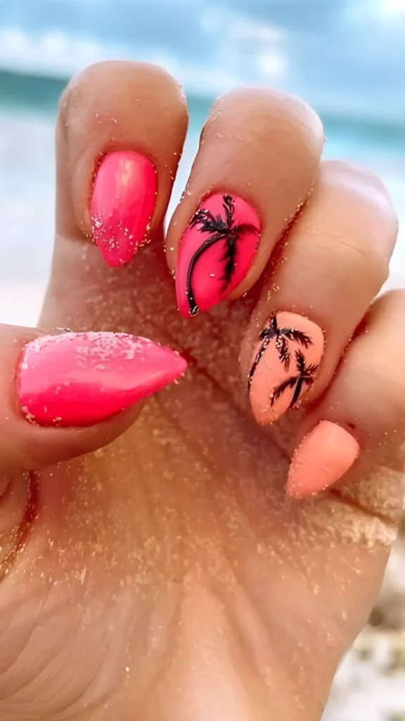 A close-up of a hand with long, stiletto-shaped nails resting on beach sand. The nails are painted in an alternating gradient of bright hot pink and soft peach, featuring delicate hand-painted black palm tree silhouettes against the tropical gradient colors.