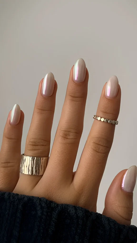 A minimalist manicure on short almond nails featuring a sheer, milky-nude base with a subtle pink-to-gold shifting iridescent glaze. The hand is accessorized with a wide, textured silver thumb ring and a delicate gold ring.