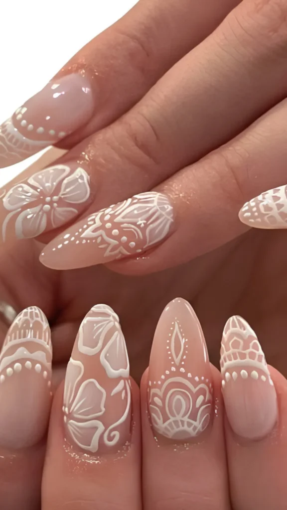 A close-up of almond-shaped bridal nails featuring intricate, hand-painted white lace and floral mandalas over a sheer nude base coat, creating a delicate henna-inspired wedding look.