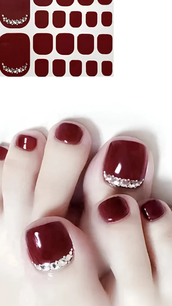 A two-panel image featuring a luxury deep burgundy pedicure. The top portion shows a sheet of solid dark red nail wrap stickers with pre-arranged rhinestone borders. The bottom portion shows the applied pedicure on feet, featuring a high-gloss wine-red color beautifully accented by a crescent curve of glittering silver rhinestones along the cuticles of the big toes.