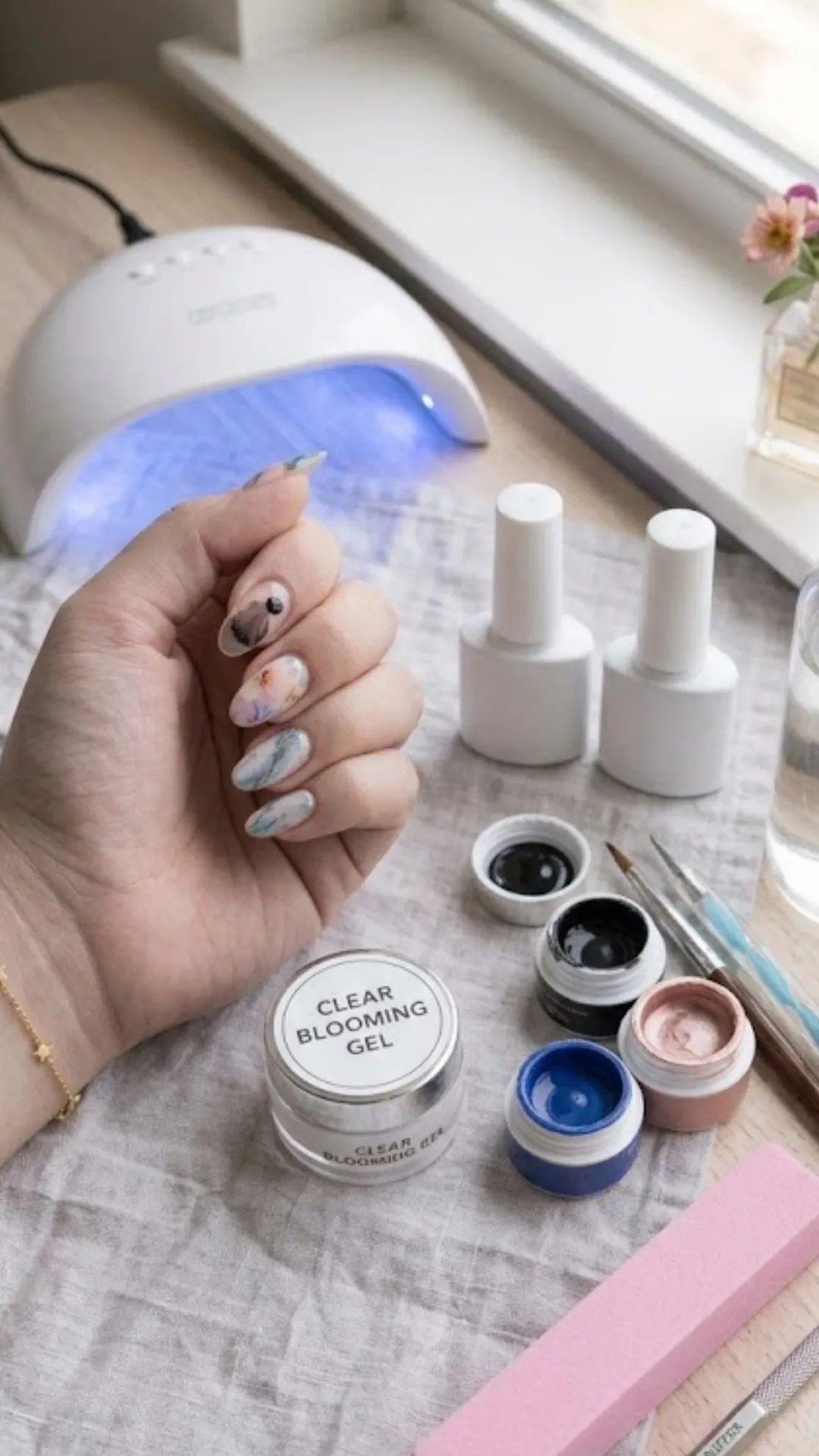 A flat lay workspace prepared for a blooming gel tutorial, showcasing a hand with completed watercolor floral nails next to an active white UV/LED nail lamp, small pots of colorful gel paints, detail brushes, and a prominent jar labeled "Clear Blooming Gel."