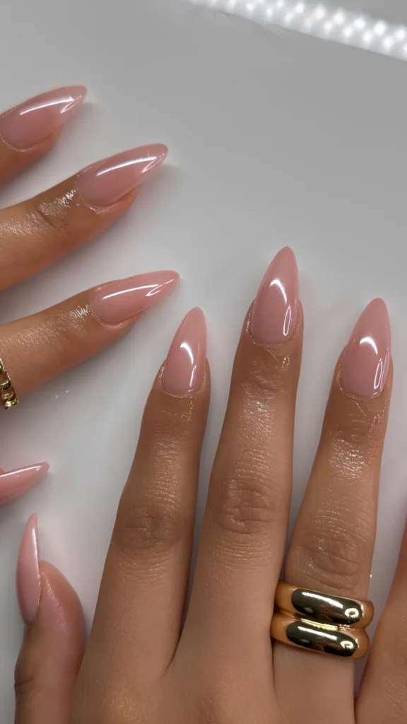 A hand with long, elegant stiletto nails painted in a glossy, semi-sheer baby pink lacquer. The skin is oiled and glowing, and the middle finger is accessorized with a thick, smooth gold band ring.