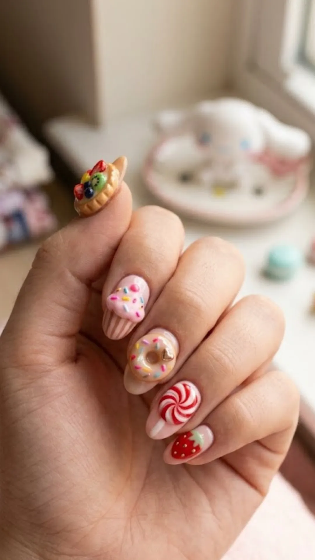 Playful 3D dessert-themed nail art featuring miniature charms of a fruit tart, a pink frosted cupcake with sprinkles, a glazed donut, a red swirl peppermint candy, and a painted strawberry on the pinky nail.