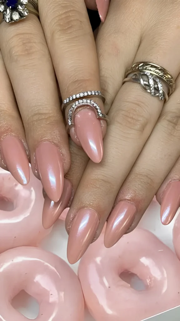 A close-up of fingers decorated with long, oval-shaped mauve-pink nails finished with a striking silvery-blue pearlescent chrome powder. The fingers are adorned with multiple textured silver bands and set against a background of glossy, pink-glazed donuts.