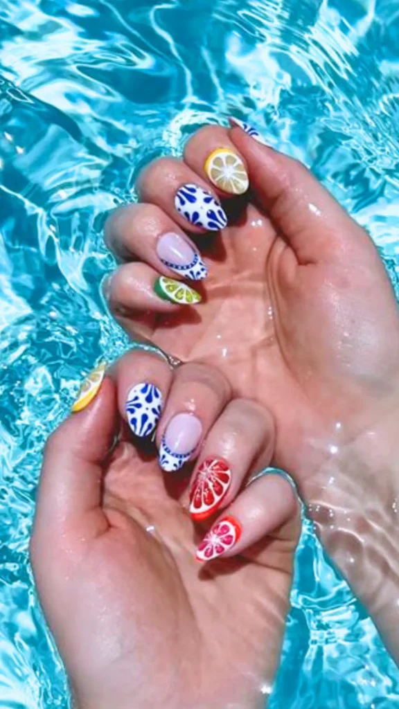 Two hands submersed in clear, rippling turquoise pool water, showcasing a playful summer manicure. The oval nails are decorated with mixed Mediterranean-style designs, including bright yellow lemons, red grapefruits, and blue-and-white tiles.