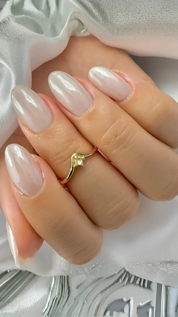 A close-up of a hand showcasing well-groomed, long oval nails with a clean, milky white base coat, topped with a luminous, high-shine pearlescent glaze. A delicate chevron-shaped gold ring sits on the ring finger.