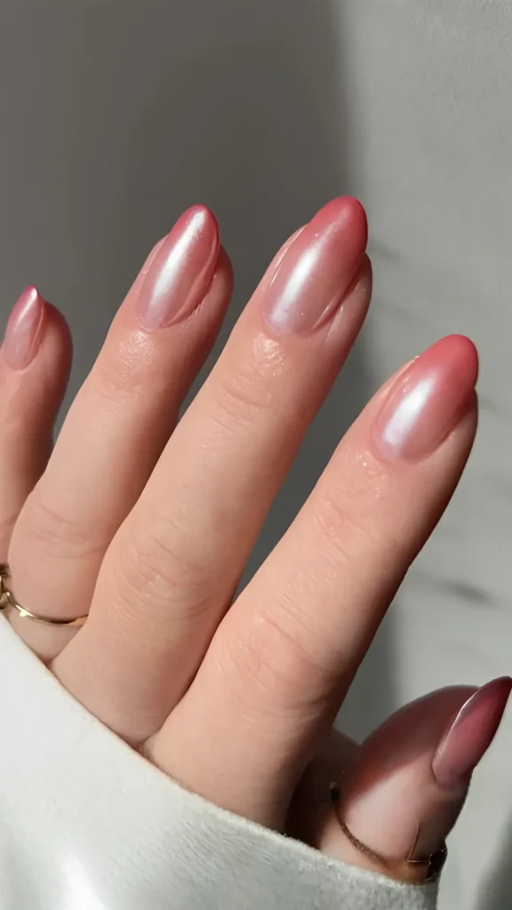 A hand showcasing an ombré gradient manicure on oval nails, transitioning seamlessly from a warm coral-rose tone at the tips to a cooler, translucent pink near the cuticles, enhanced by a frosty white glazed finish.