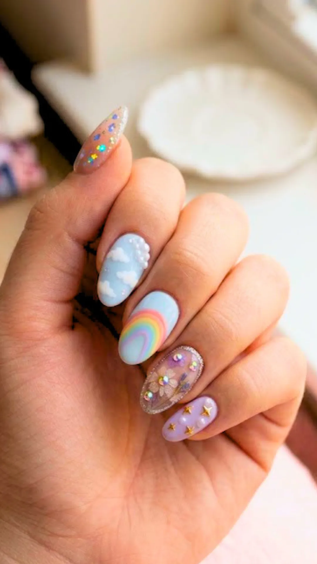 An elegant variation of sky-themed nail art with iridescent glitter on the thumb, 3D pearl clusters over white clouds, a pastel rainbow, a clear nail encapsulated with pressed flowers and rhinestones, and gold stars over lavender.