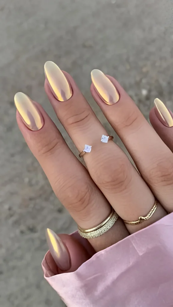 A hand with long, slender almond nails featuring a glamorous, warm champagne-gold metallic chrome finish that gleams under the light. The hand is styled with minimalist gold jewelry against a soft pink fabric cuff.