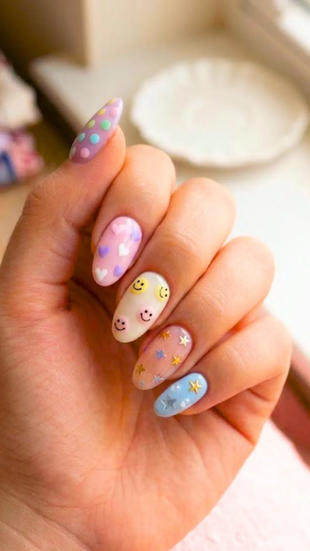 Colorful and whimsical kawaii nail art showcasing pastel green and pink polka dots, 3D pastel hearts on pink, retro colorful smiley faces on an off-white base, and tiny star charms scattered across clear and baby blue nails.
