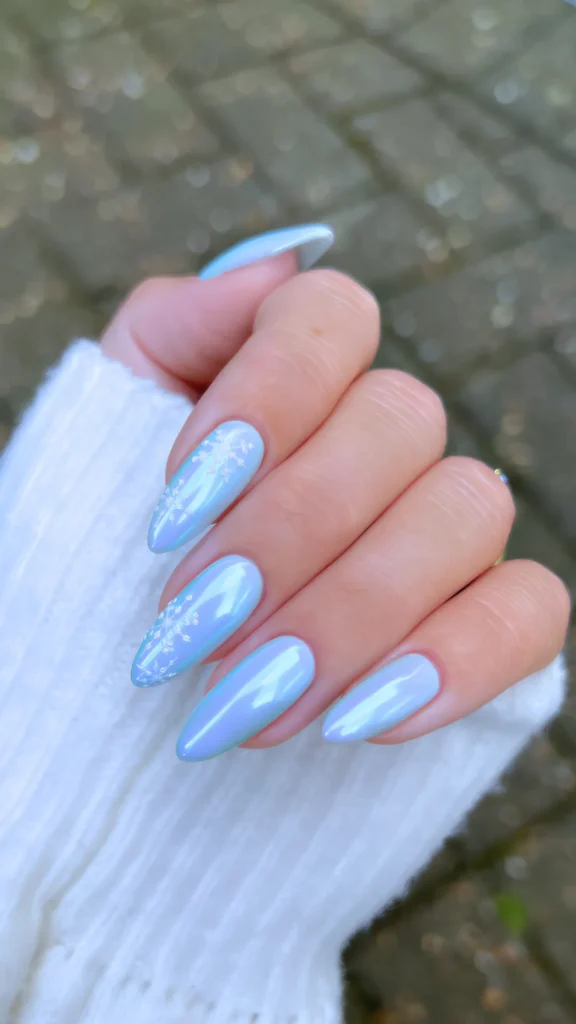 A winter-themed manicure on long oval nails with a frosty, ice-blue base coat and a shimmering glaze overlay, decorated with delicate white hand-painted snowflake details on the index and middle fingers.