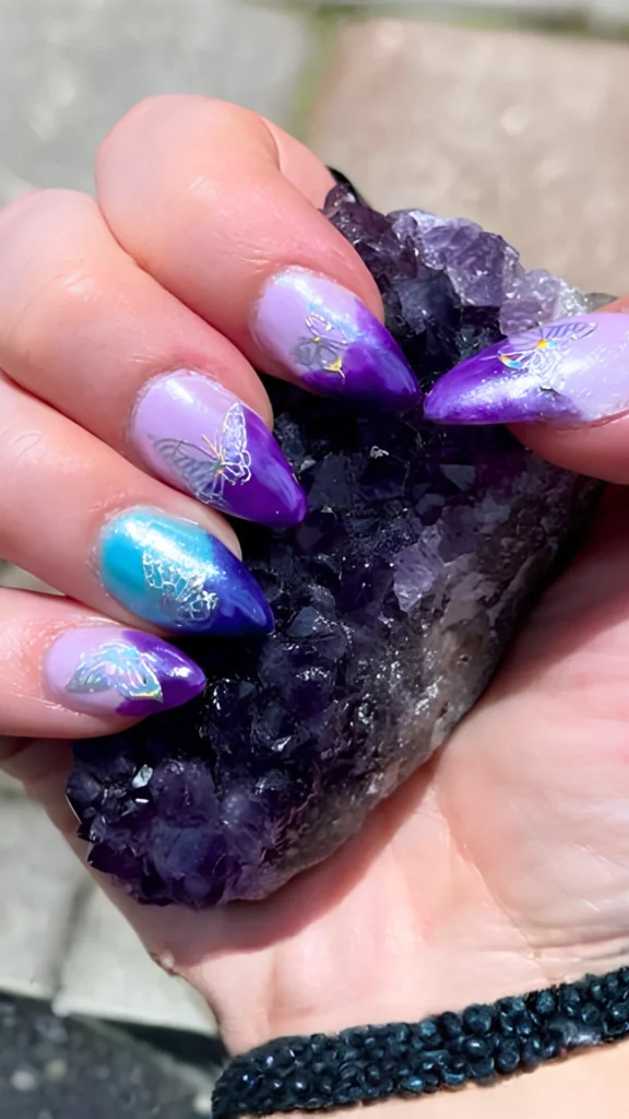A bold and colorful manicure featuring almond-shaped nails with a vibrant purple, magenta, and blue gradient. Each nail is accented with a shimmering, iridescent butterfly decal while the hand cradles a large, raw purple amethyst crystal cluster.
