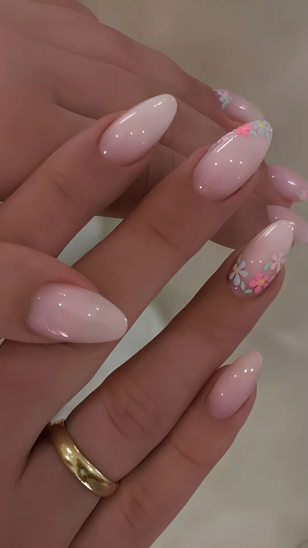 A close-up view of a hand with elegant, long oval-shaped nails showcasing a chic and feminine manicure. The nails feature a glossy, semi-sheer milky pink base coat. The ring finger stands out as an accent nail, decorated with a delicate arrangement of small, hand-painted pastel flowers in shades of pink, white, and soft blue, complete with tiny green leaves near the tip. A simple gold ring is visible on the pinky finger.