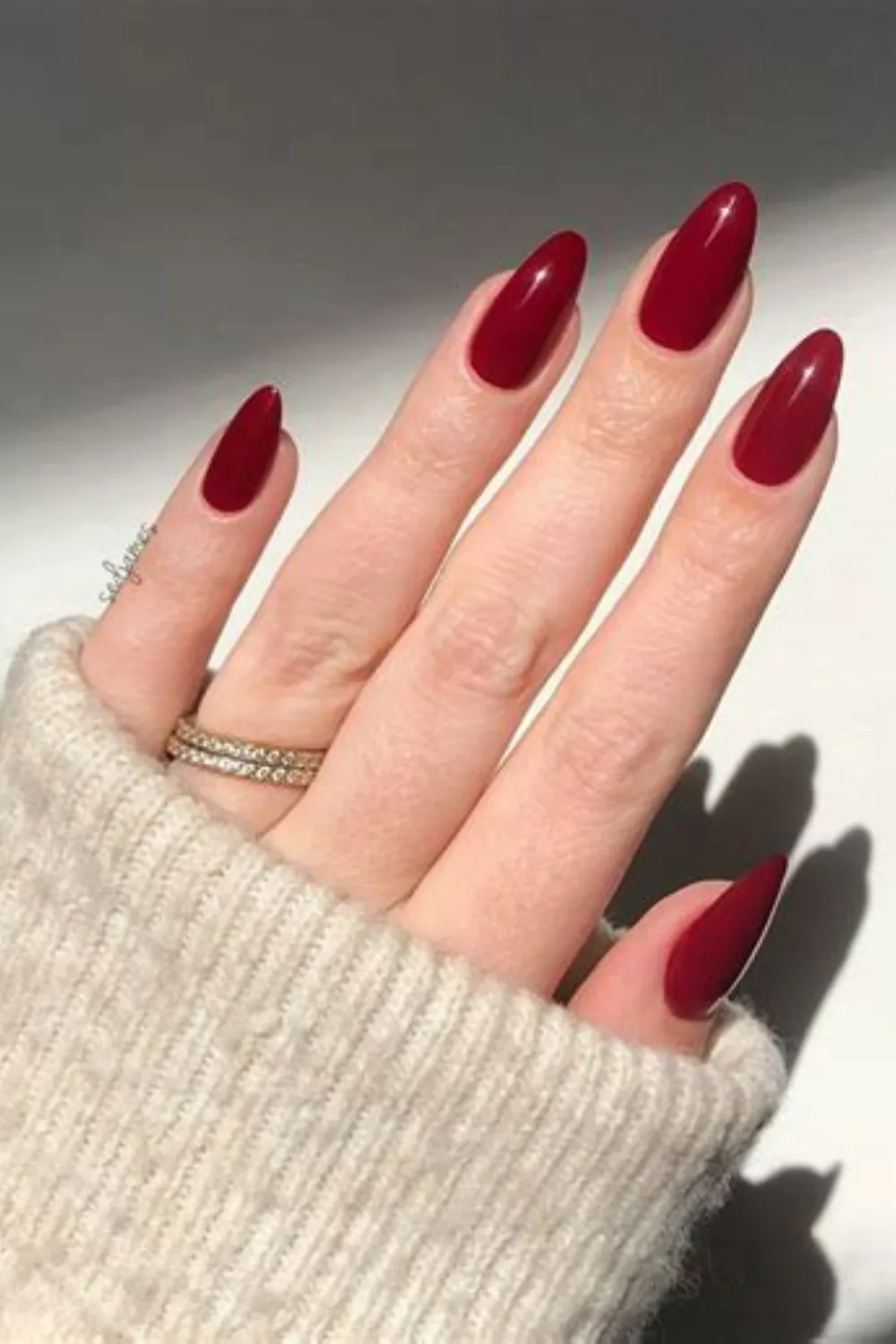 A close-up photograph showcasing a powerful and classic solid red manicure on long, oval-shaped nails. The fingernails feature a high-gloss, glossy cream finish in a rich, deep crimson red. The model's hand is partially covered by the cozy sleeve of a cream-colored knit sweater, and a delicate gold band encrusted with tiny crystals is visible on the ring finger under bright, directional sunlight.