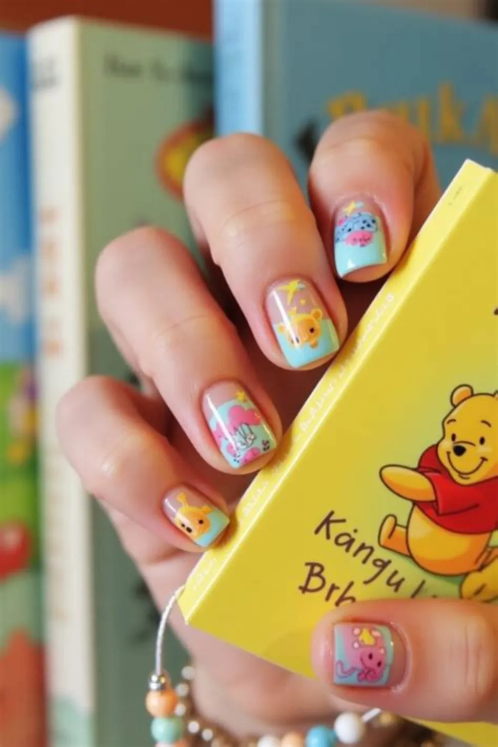 A close-up photograph showcasing a cute Winnie the Pooh-themed baby shower manicure on short, squared nails. The nail design features pastel sky-blue and soft pink background panels decorated with hand-painted characters, including Winnie the Pooh, Piglet, and Tigger under yellow stars. The model holds a bright yellow children's storybook featuring Winnie the Pooh on the cover, perfectly matching the nursery-themed nail art.