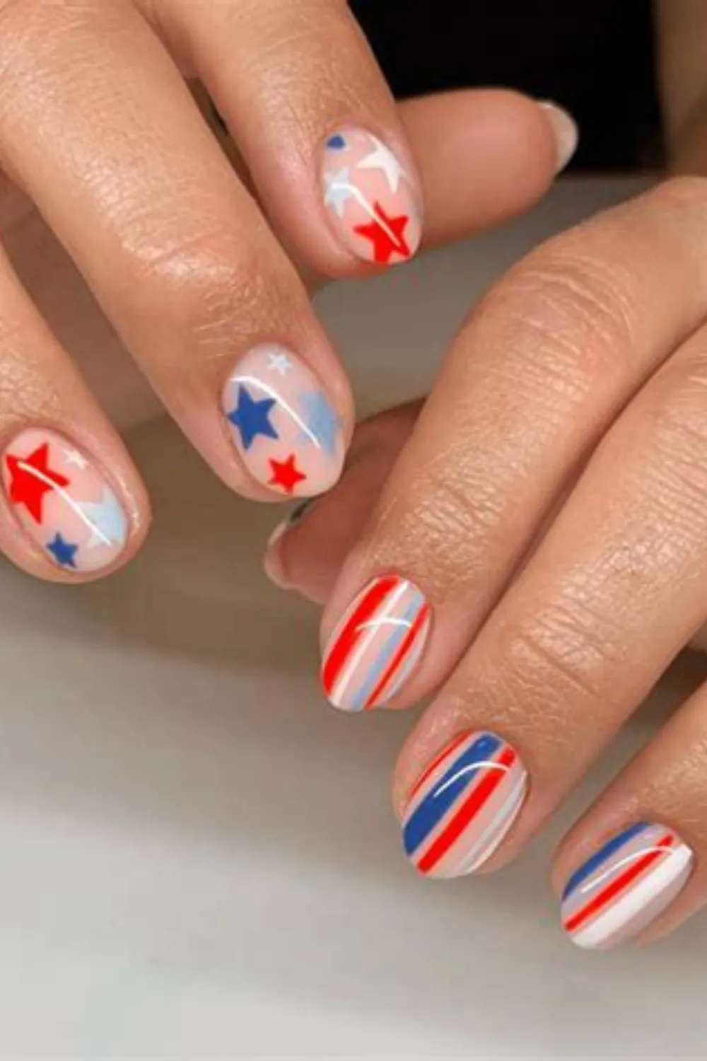 A detailed close-up shot of two hands displaying a festive 4th of July manicure on short, rounded nails. The left hand features a sheer, glossy nude base decorated with scattered hand-painted red, white, and blue stars. The right hand showcases an alternating patriotic pattern of thin, clean diagonal stripes in red, white, and blue polish over a natural, high-gloss base coat.