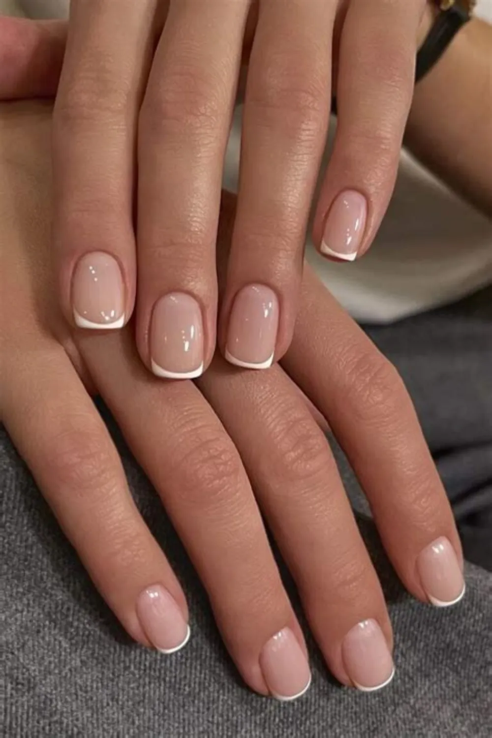 A close-up photograph showcasing a clean, classic micro-French manicure on short, neatly squared nails. The nail beds are coated in a sheer, glossy natural pinkish-nude gel polish. The tips feature ultra-thin, precise hand-painted smiles of crisp white polish, creating a refined, timeless, and understated aesthetic suitable for any formal or casual setting.