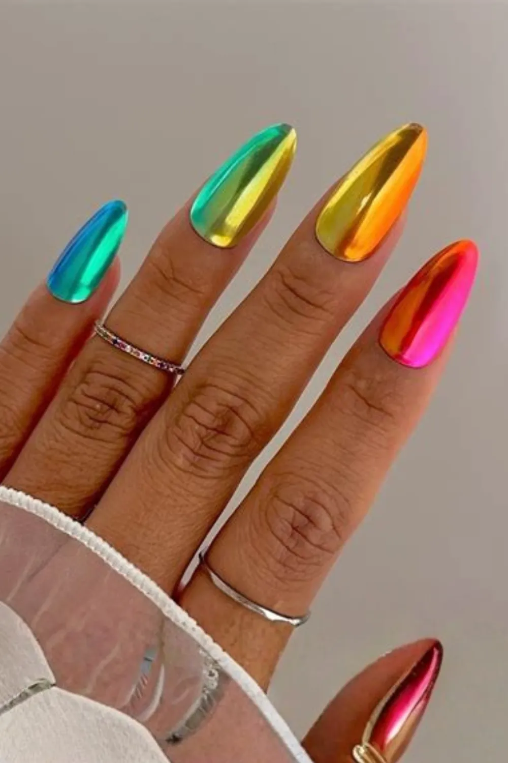A close-up photograph showcasing a stunning rainbow metallic manicure on long, stiletto-shaped nails. Each nail features a ultra-reflective, liquid chrome finish in a different bold shade, including electric cyan, neon green, bright yellow, radiant orange, and vivid magenta pink. The hand is accessorized with delicate thin gold rings, including one encrusted with tiny rainbow-colored gemstones.