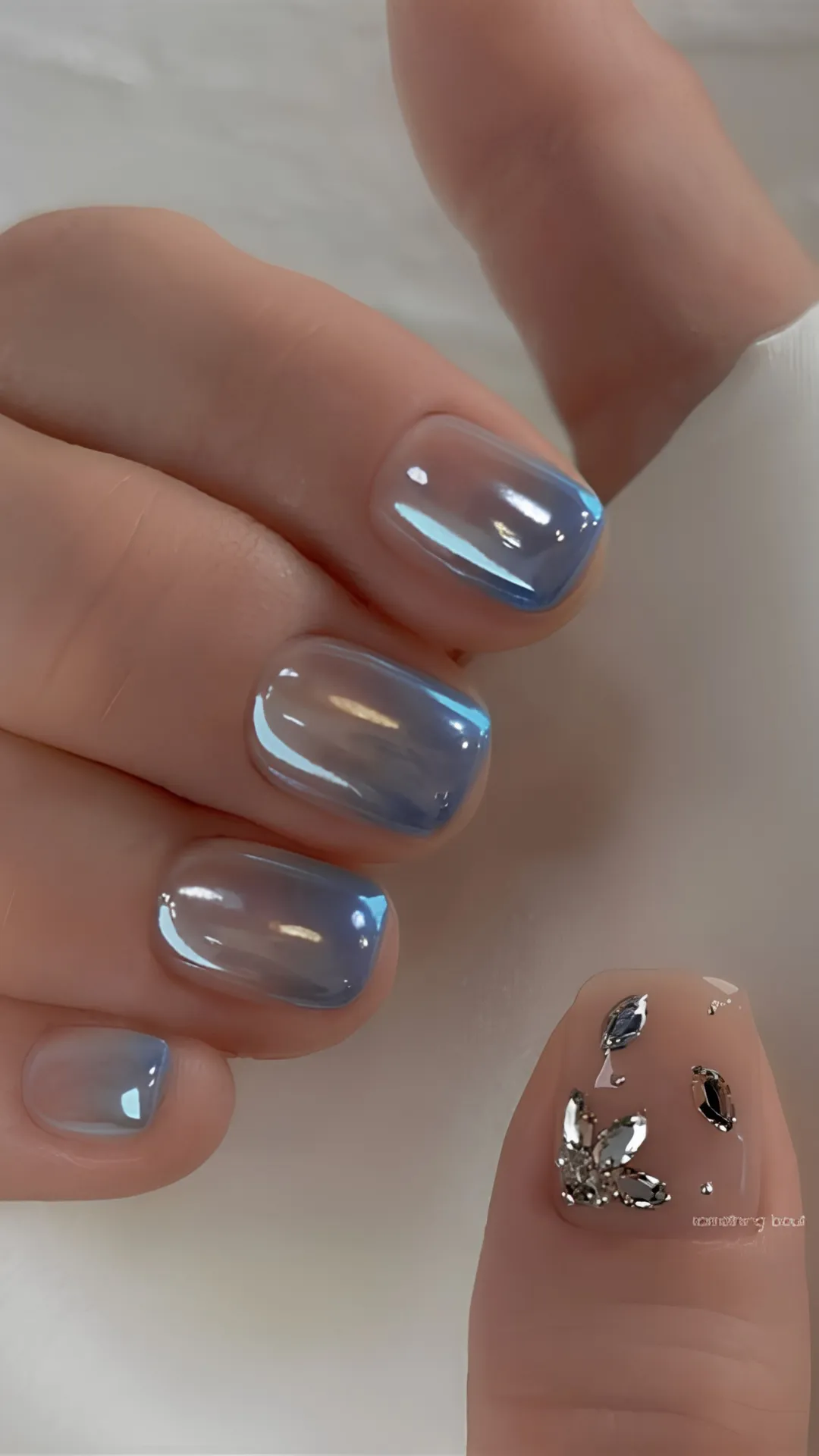 Close-up shot of a hand showcasing a modern Korean manicured look. The short, square-shaped fingernails feature a luminous, metallic ice-blue chrome finish with an iridescent sheen that catches the light. The thumb stands out with a neutral, glossy beige base adorned with an intricate 3D arrangement of clear and reflective silver rhinestones shaped like small leaves or butterfly wings.