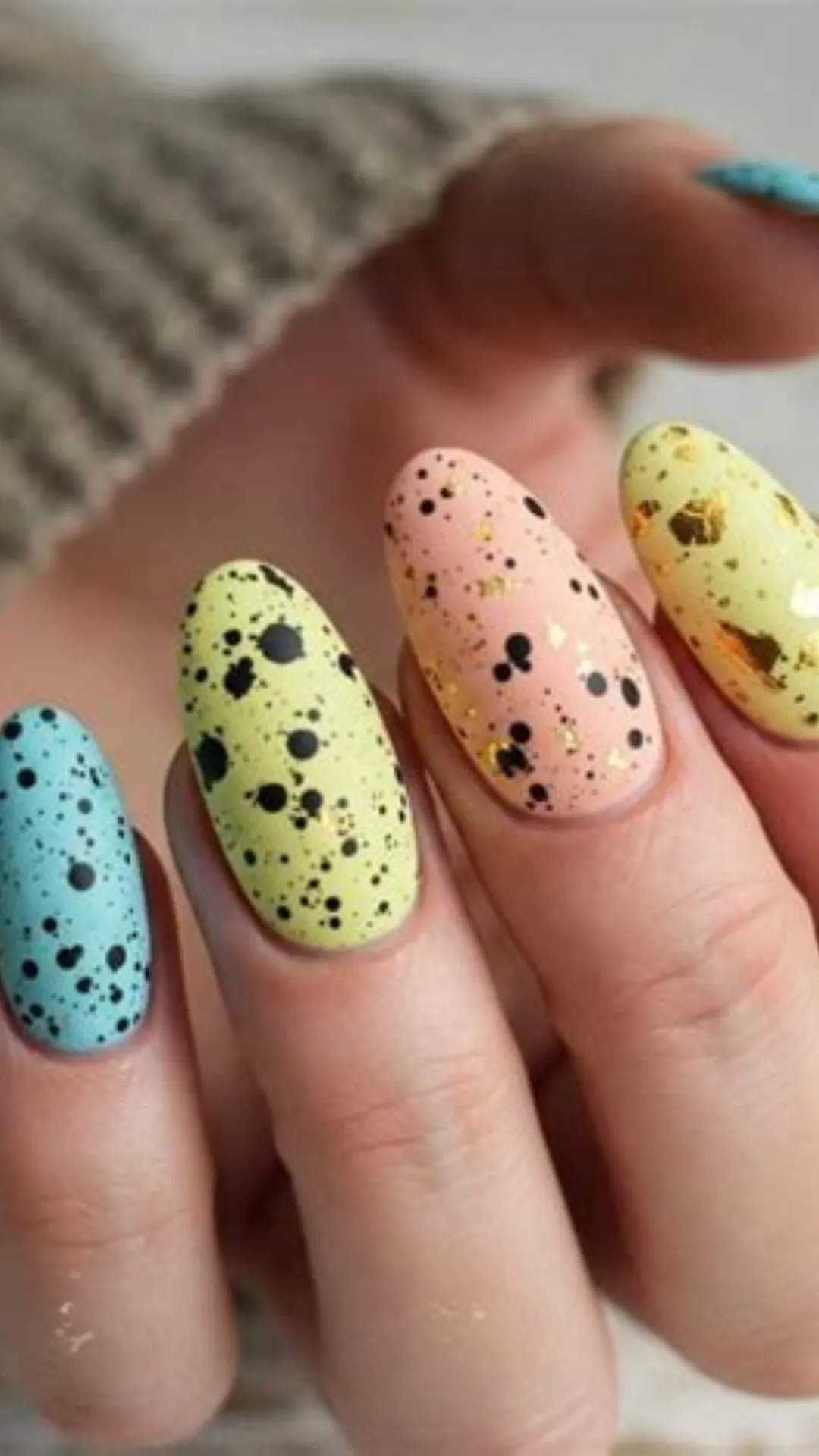 A close-up view of oval-shaped nails showcasing a creative speckled Easter egg design. Each fingernail is painted a different soft pastel color, including baby blue, light mint green, and pale peach. The matte-finished nails are covered in realistic fine black paint splatters mimicking a bird's egg shell, with some nails accented by delicate flakes of reflective gold foil that add a touch of elegance.