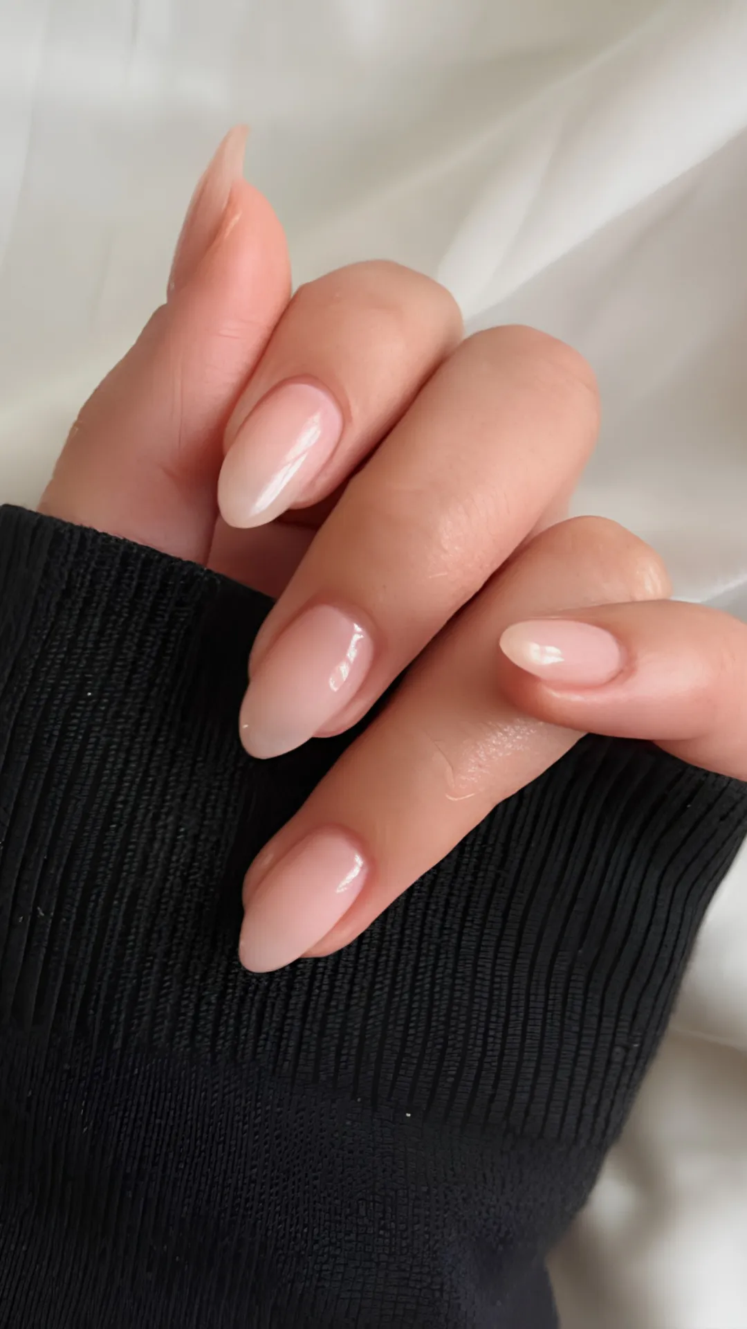 A close-up photograph displays a hand with immaculate, short almond-shaped nails. The nails are painted in a high-gloss, semi-sheer milky pink or nude gel polish, creating a natural and understated "your nails but better" look. The hand rests against a textured black knitted sleeve, contrasting with the soft, flowing white silk background. The lighting is soft and natural, emphasizing the flawless, reflective finish and clean cuticle lines. This elegant and versatile manicure is ideal for achieving a sophisticated, minimal, and everyday look with almond-shaped nails.