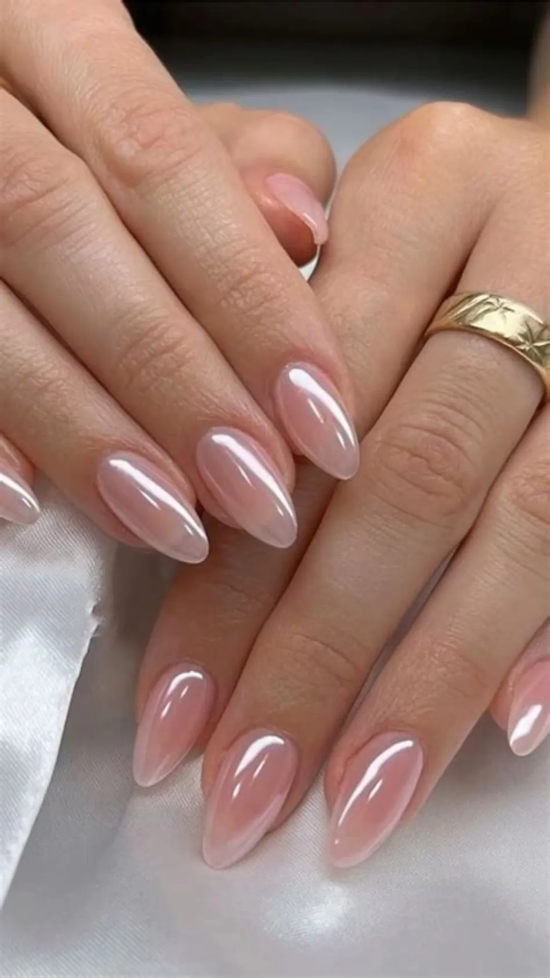 A close-up view of two hands resting together on a white satin fabric, displaying a luxurious glazed donut manicure. The long, almond-shaped nails are finished with a soft pink chrome powder overlay that creates a luminous, pearlescent sheen reflecting bright white lines of light. A thick, wide gold band with subtle star-like engravings is worn on the ring finger.